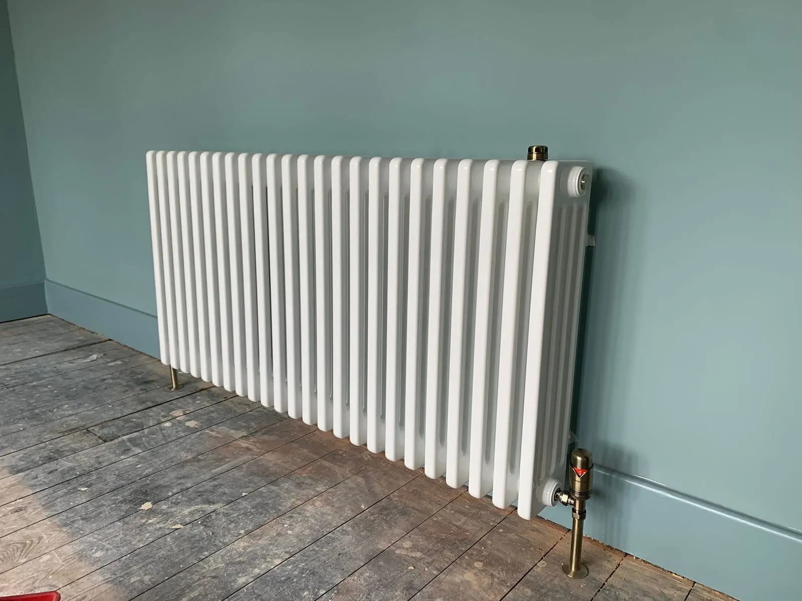 A white column radiator fitted against a teal wall above bare floorboards
