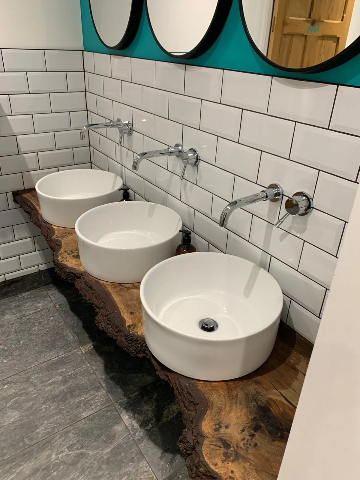 Three counter-top basins on a live-edge timber shelf against white metro tiling in a commercial washroom